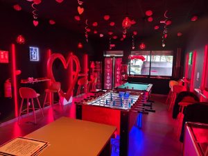Vista interior de un bar, donde se ven mesas, sillas y un futbolín